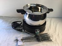 Stainless steel fondue pot resting on a round black electric heating base with temperature dial and power cord attached. Eight fondue forks with black handles laid in front, bundled in white cardboard sleeve.