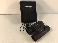 New Tasco 12x8mm compact binoculars shown with the included black belt case on a flat surface, front angle with logo visible.