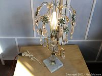 Full view of vintage crystal table lamp with brass flowers and crystal prisms illuminated, showing marble base and cord.