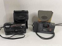 Two 1990s compact film cameras with their carrying cases placed on a flat surface. Left is a black Nikon EF100 and right is a Kodak Ektralite camera with an attached wrist strap.