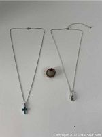 Photo of two necklaces laid flat, one with a blue cross pendant and the other with a green teardrop-shaped pendant with extender chain, next to a ring with a round central stone and elastic band.