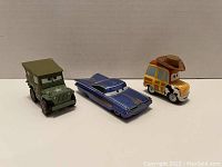 Photo showing three metal Disney Pixar Cars toys including a green army jeep, blue classic car, and yellow woodie with cowboy hat, displayed on a flat surface with a neutral background.