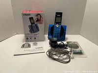 VTech blue cordless phone system with base, handset on base, power adapter, phone cord, additional accessories, user manuals and original box visible