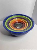 Photo of seven nested colorful mixing bowls sealed in plastic wrapping, varying sizes from largest blue outer to smallest red inner bowl.