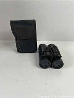 Photo of folded black Bushmaster binoculars placed next to their black fabric carrying case with flap closure.