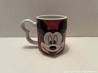 Front view showing large 3D raised Mickey Mouse face on pink mug with Mickey-shaped handle.