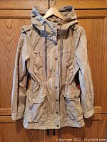 Front view of tan women's parka jacket hanging on hanger showing hood, full button and zipper front closure and front flap pockets.