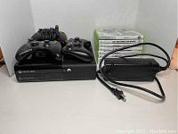 Front view of Xbox 360 console with three controllers on top, power adapter next to it, and a stack of Xbox 360 game cases visible.