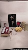 Photo showing assortment of Asian decor items including mugs, ashtray, pendant, framed thimbles, and peace wall hanging