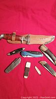 All eight cutting tools arranged on a red background, including seven folding knives and one fixed blade knife with leather sheath, showing varying wear and rust.
