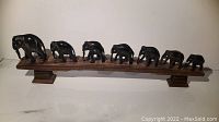 Front view of seven black hand-carved elephant figurines arranged in a row on a wooden display stand with two pedestal feet.