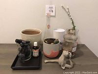Photo showing beige ceramic pot, small black tray, candle in glass jar, white yoga frog figurine on wooden table with some other containers and plants.