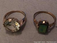 Pair of rings side by side: left ring 10K gold with large clear stone, right ring gold tone with green stone