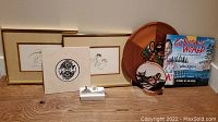 Front view of two framed bird prints, Coast Salish salmon people art print, wooden eagle tray, ceramic plate, children's book and candle arranged on wood surface