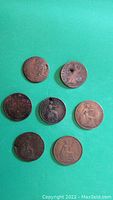 Overview of 7 old British coins on green surface, showing varying degrees of wear and corrosion, including 2 coins with holes.