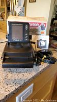 Front angled view of the black SupraPro I.D. Portable fish finder device resting on its folding stand on a countertop, original box and instruction manual behind it.