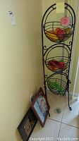 Full view of the black metal 3-tier basket stand with artificial fruit in baskets. Two framed wall art pieces and a small decorative item on the floor beside stand.