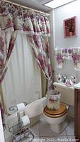 Full view of bathroom decor installed showing floral shower curtain with valance, plastic liner, matching hand towels on rack, waste basket with fabric cover, toilet seat cover, metal toilet paper/magazine holder, soap and lotion dispensers on tank, and wall floral art.