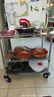 Photo of metal cart with three shelves filled with various kitchen items including copper pans, stainless steel pots, blender, and mixer.