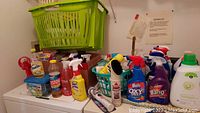 Cleaning supplies lot with green laundry baskets, red dish drainer on top various spray bottles and detergent bottles arranged next to each other on a white surface.