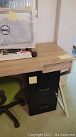 Desk with wood-grain top, Dell monitor box on desk, black metal file cabinet tucked underneath, and green/black office chair visible