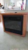 Front view of medium oak wooden cabinet housing electric fireplace insert with black front frame and remote control on top.