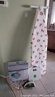 Full view of ironing board with floral cover, foot spa box, white digital scale, and electric iron on tiled floor.