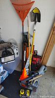 Image showing long-handled orange leaf rake, yellow Stanley shovel, Earthwise cordless blower, and other assorted tools and boxes on the floor against a wall.