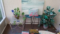 Photo showing metal side table with a plastic tiled top, two metal-legged stools with flamingo themed pillow on one, ocean flamingo wall decor, live and artificial plants, and boxed board games displayed on floor