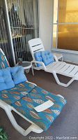 Two white plastic patio lounge chairs; one with blue patterned cushion and blue tufted pillow, the other with solid blue tufted pillow, positioned on patio with sliding door background.