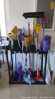 Photo showing brooms, mops, bucket with cleaning liquids, and a black plastic stand holding the items in an upright position.