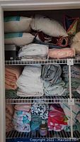 Shelving unit with multiple pillows, folded towels, blankets and bed sheets in mixed colors and patterns.
