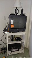 Black CRT TV on white stand with remote and various electronics