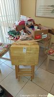 Four wooden TV trays stacked with folding legs visible, placed in front of a table holding boxes and plastic containers.