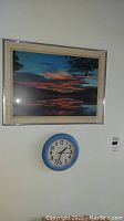 Framed sunset wall art and blue rim wall clock mounted below it on a white wall for decorative use.