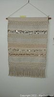 Woven wall hanging with horizontal sequin stripes and fiber fringe hanging on a wall.