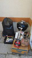 Weber charcoal kettle grill, grill cover, boxed utensils, charcoal briquettes, and other grilling supplies arranged on ground.