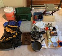 Photo of assorted camping and travelling gear including backpack, pots and pans, water cooler, foldable seat, and miscellaneous kitchen and camping items spread on the floor.