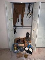 Carhartt overalls hanging inside closet with boots, helmet, knee pads, gloves, and small bins of miscellaneous tools and materials.