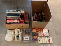 Photo showing two large boxes containing multiple board games stacked and organized. One box holds various boxed games including Animalia, Scrabble, and more, along with a cloth doll and card games labeled Mr. M and Mr. Uppity. Another box contains books and additional games.