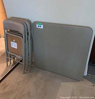 Set of one folding beige table and four matching folding chairs stacked beside it.