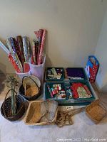 Photo showing rolls of various decorative gift wrapping paper in a plastic bin, three green trays with assorted ribbons and gift tags, and seven different woven baskets on the floor.