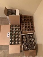 Four cardboard boxes containing multiple clear glass tumblers each separated by cardboard dividers for protection. The glasses are cylindrical and transparent.