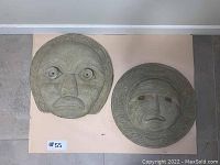 Two round concrete indigenous style mask plaques viewed from above, showing detailed carved facial features and decorative border patterns.