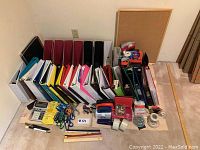 Photo showing a large assortment of office supplies and organizational items including many binders in various colors, magazine holders, cork board, scissors, staplers, and other desktop accessories arranged on a floor.