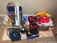 Photo showing assortment of outdoor-related items including tennis racket, cycling pump, helmet, kites, and cycling accessories arranged on floor