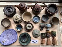 Top view of 23 assorted pottery items showing various bowls, cups, lids, and plates on a beige background.