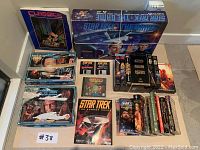 Wide shot showing various Star Trek and Star Wars boxed board games, VHS tapes, DVDs, books, and a Warcraft CD all laid out on the floor. Boxes show wear and tape residue.