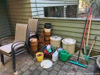 Wide view showing 6 patio chairs, stacked terracotta pots, two large ceramic Imperial pots with lids, garden hose, watering cans, and several gardening tools including rakes and hoes.