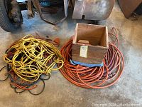 Overall view of large pile of yellow electrical extension cords and coiled red air hoses with wooden box on top.